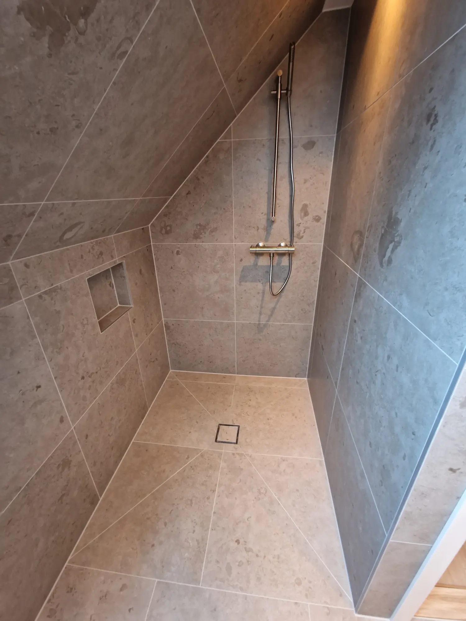 Badrumsrenovering av duschutrymme under snedtak, kaklat med stora gråa plattor, optimerar ytan effektivt.