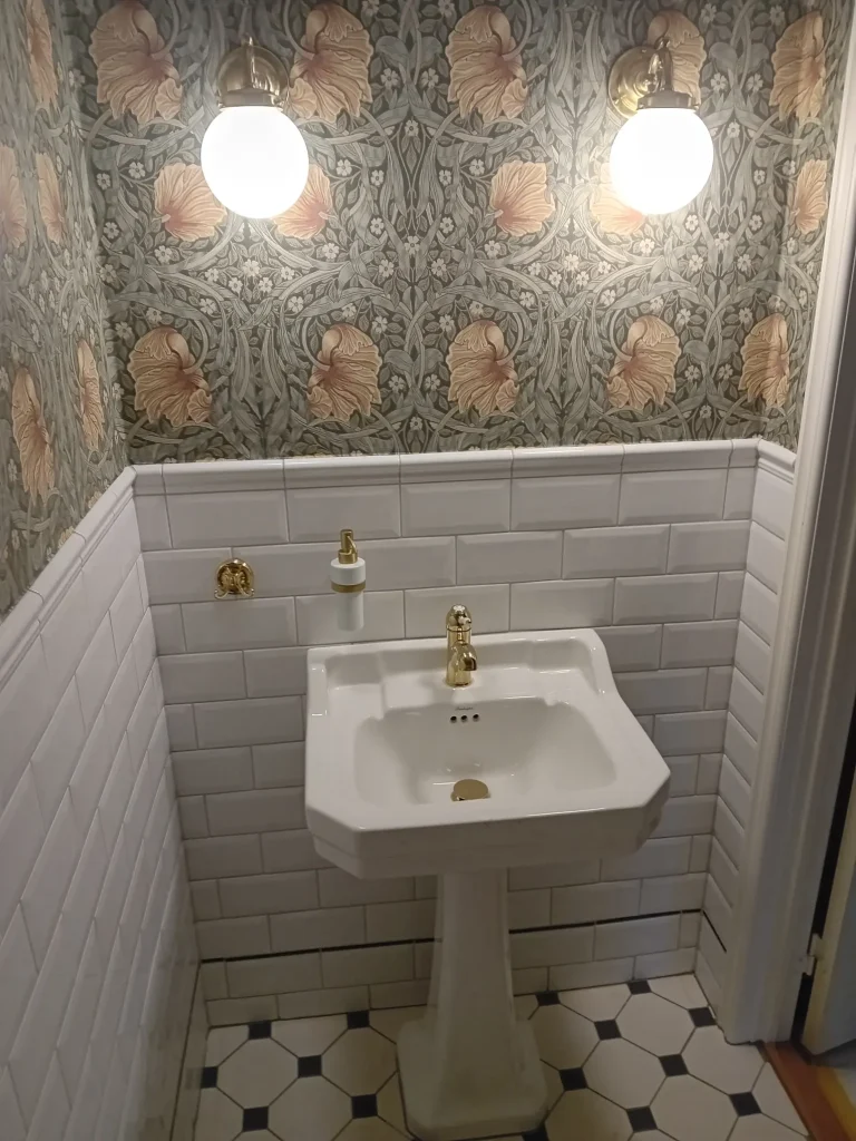 Stilren badrumsrenovering med klassisk tapet, mjuk belysning och exklusiva gulddetaljer.