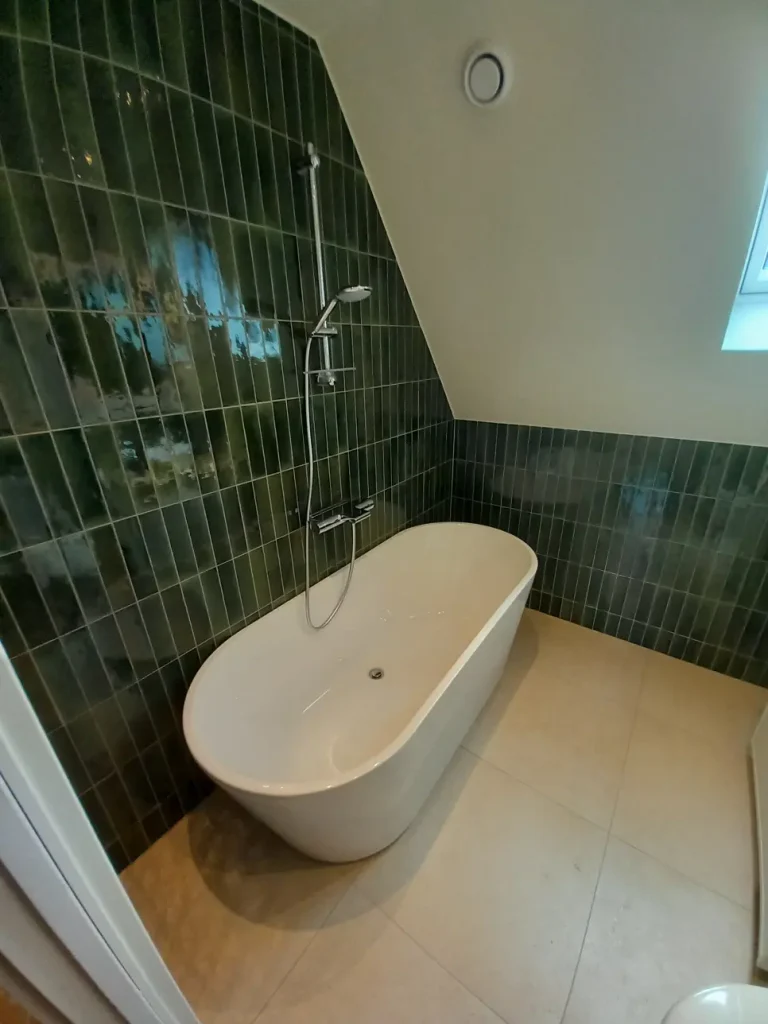 Badrumsrenovering med fristående badkar under snedtak, omgivet av grönt stående kakel och ljust golv.