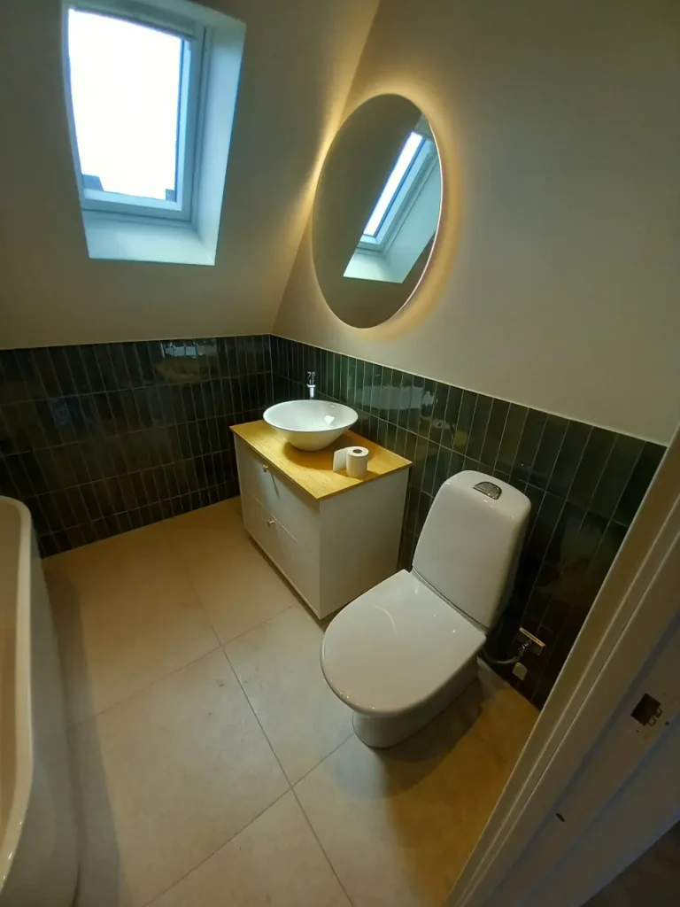 Helhetsbild av badrumsrenovering med WC, stilren kommod och ljust golv som kontrasterar mot grönt stående kakel.