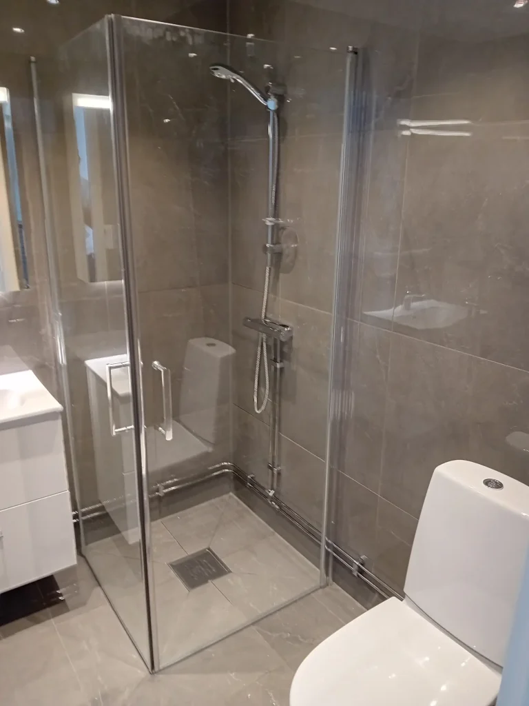 Badrumsrenovering med glaskabin för dusch och modern toalett, gråa 60x60 cm plattor för ett enhetligt uttryck.